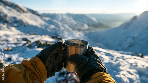 Gloved hands holding a steaming metal cup against a sunlit snowy mountain valley with rugged peaks and winter alpine landscape in soft focus