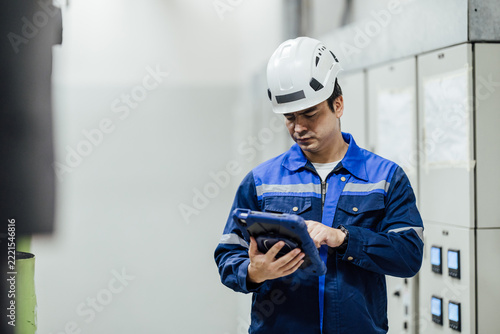 Industrial technician in hard hat using a tablet to inspect electrical control panels inside a factory, emphasizing safety, maintenance, and digital operations.