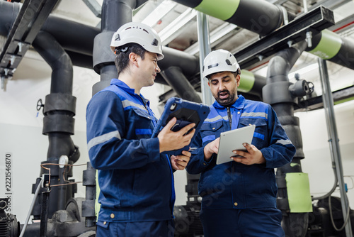 Industrial engineers in protective gear inspecting machinery with digital tablets in a mechanical room with large pipes and valves.