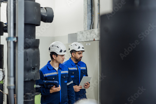Two industrial technicians in blue uniforms and helmets inspect machinery in a plant room, using a tablet and notes during a safety maintenance check.