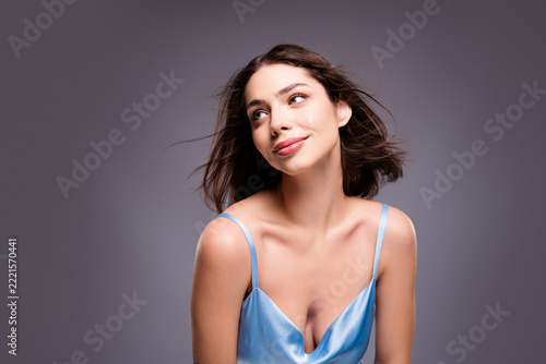 Young woman in blue satin dress smiling and looking up in studio portrait, natural makeup and flowing hair conveying confidence and joy