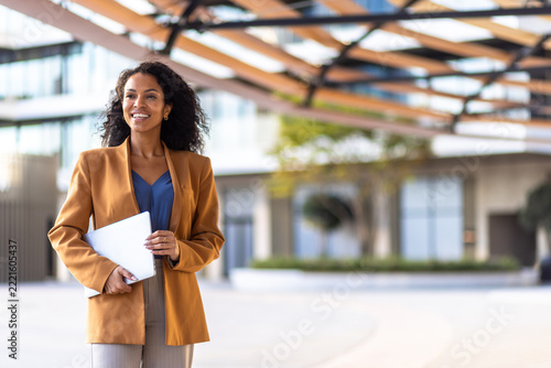 Confident african american businesswoman holding laptop, smiling while standing outside office building, representing career success and entrepreneurship
