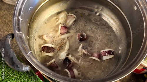 Close up shot of fresh squid rings and tentacles boiling in hot savory broth inside a metal pot at a Thai street food market stall.