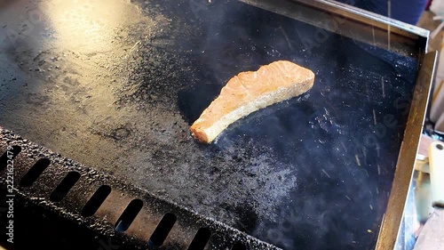 Close up shot of a delicious hot salmon fish steak frying with smoke and steam on a flat iron griddle at a Thailand street food market.