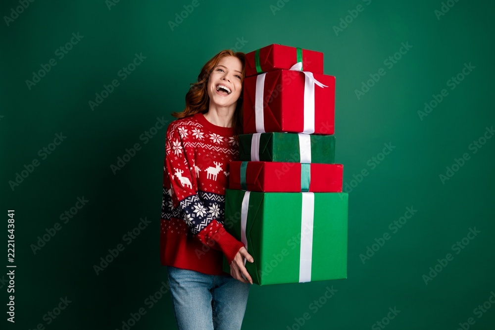 Naklejka premium Happy young woman in red Christmas sweater carrying a tall stack of wrapped gifts against a green background