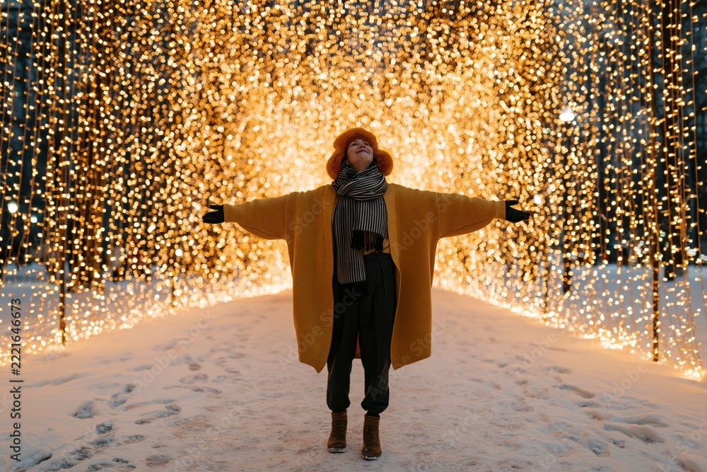 Fototapeta premium Person with outstretched arms in illuminated holiday light tunnel on snow for winter holiday background and celebration banner with radiant golden bokeh