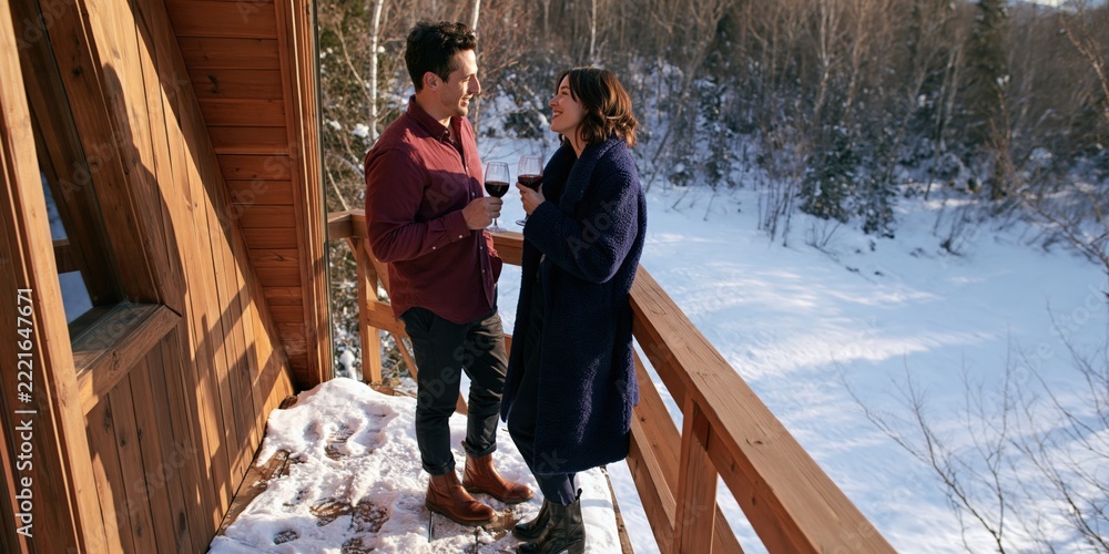 Fototapeta premium Couple drinking red wine on a snowy cabin balcony overlooking forest for romantic winter getaway banner background