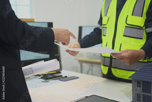 Worker and colleague examine blueprint in office planning, with paperwork spread across desk for discussion