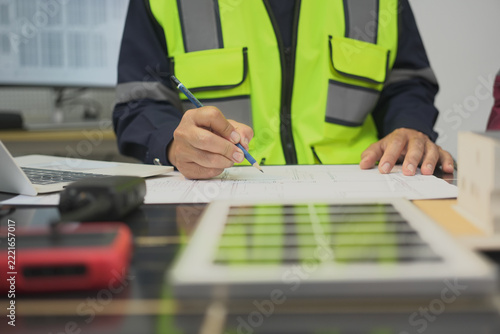 Worker drawing blueprint wearing reflective vest at office desk with construction planning document and laptop