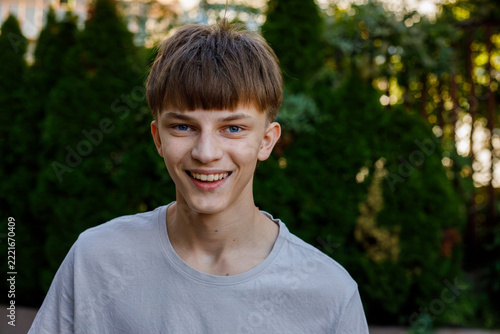 Happy teenage boy with brown hair and bangs wearing gray shirt, smiling and laughing outdoors in a green garden, cheerful lifestyle portrait of a boy, grainy film texture, high quality photo