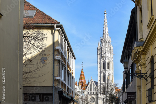 Historic street in Budapest's Buda Castle District frames Matthias Church, with colourful tiled roofs and Gothic architecture beneath a bright blue sky.