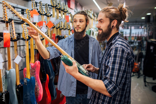 Two musicians stand together at guitar row. One young man hold guitar white another touches its bar. They are in guitar store.