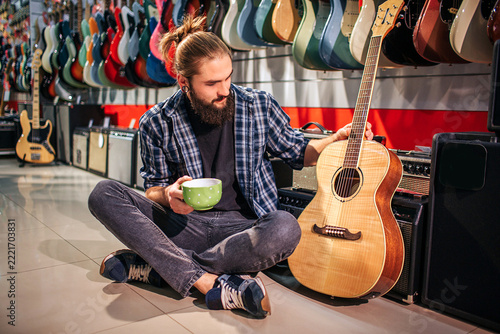Young hipster has rest by sitting on floor. He hold cup in hand. Another one touches acoustic guitar nearby him. Young man looks at instrument.