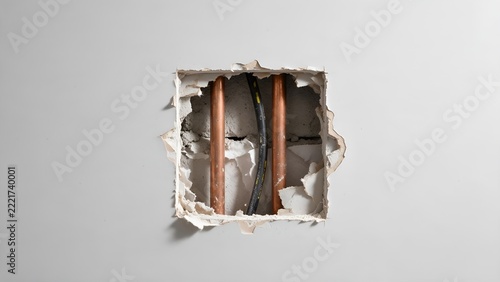 Square hole cut into a white drywall interior partition displaying exposed copper plumbing pipes and black electrical wiring during residential home renovation work