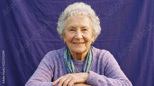Happy elderly woman with white hair smiling warmly wearing a purple sweater and scarf against a purple background