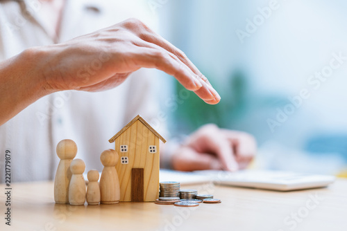 Person hands protecting miniature wooden house and family figurines with stacks of coins on table, home insurance and financial planning concept