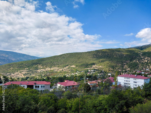 Scenic view of a small mountain town surrounded by lush green hills, vibrant forest landscapes, red-roofed buildings, and a bright blue sky with fluffy white clouds