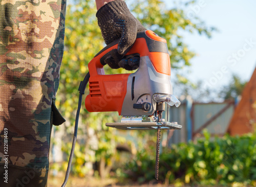 Fotografie Carpenter hand in a protective glove holding an electric jigsaw tool with a long saw blade outdoors
