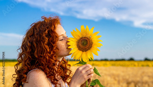 A young woman with vibrant red curly hair and freckles blissfully smells a blooming sunflower in a golden field. This peaceful summer scene evokes joy, natural beauty, and a deep connection to nature.