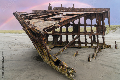The Peter Iredale, a four masted steel hulled transport ship, built in England, round aground in 1906 and remains on the shore of the Oregon Coast in the Pacific Ocean with a rainbow sky.