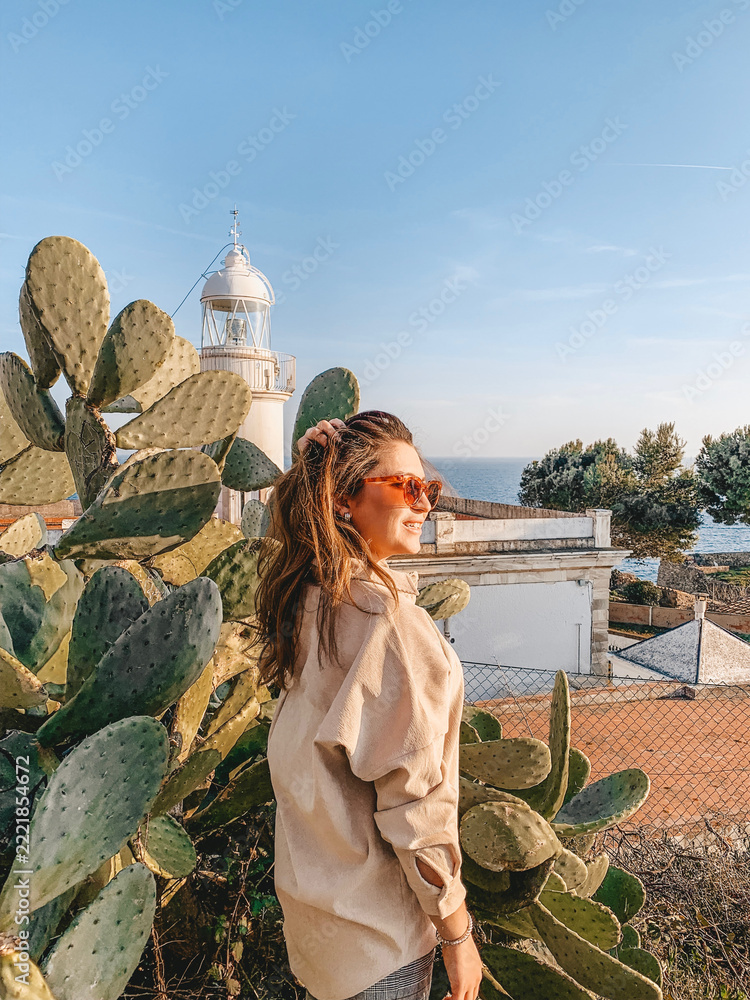 Fototapeta premium Young woman smiling beside large prickly pear cactus at coastal viewpoint with lighthouse and sea in warm golden hour light, scenic summer travel portrait by Mediterranean shore