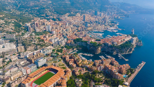 Aerial view of the principality of Monaco, featuring the Stade Louis II, dense urban architecture, the Mediterranean coastline, and the busy harbor with yachts, Monaco.