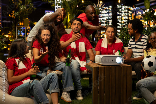 Fotografie Football fans react passionately while watching a decisive soccer match projected onto a screen on a rooftop terrace, shouting during a tense live sports broadcast with fellow supporters