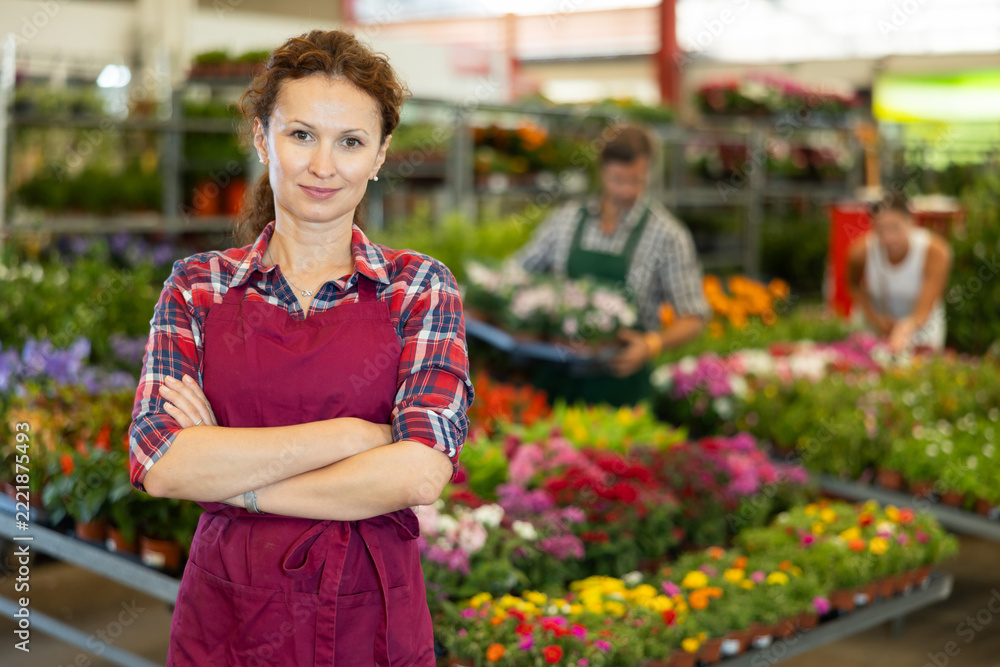 Fototapeta premium Woman worker stands in aisle between rows with variety of flowering plants in pots, waiting for visitors to consult clients. Floral supermarket, assortment of potted ornamental plants