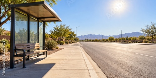 Modern bus stop shelter with empty bench on sunny suburban street. Wide sidewalk and road with desert mountains in background. Summer heat and shade concept. Banner, copy space
