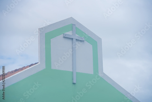 Christian Cross on Church Facade: A simple, modern Christian cross stands prominently on the light green facade of a church building, set against a softly clouded sky.