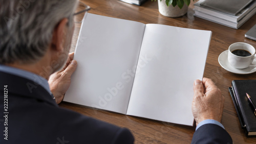 Senior businessman holding an open blank book at a wooden desk with coffee and office accessories