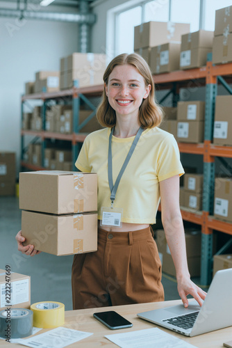 Portrait of smiling young adult Caucasian female holding cardboard packages near laptop in warehouse. Useful for ecommerce logistics, supply chain management, vertical
