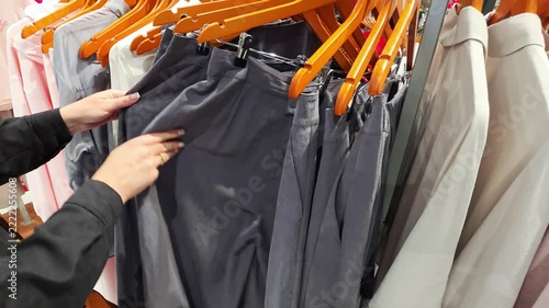 Hands of woman inspecting fabric of dark grey trousers hanging on wooden hangers in clothing store, shopping for new apparel on retail display rack in boutique