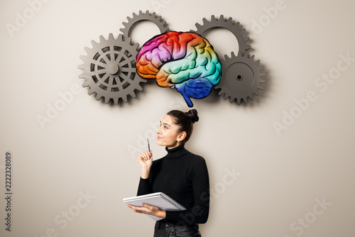 Businesswoman with notebook beneath colorful brain and gears, symbolizing education, brainstorming, learning, intelligence, and creative thinking.