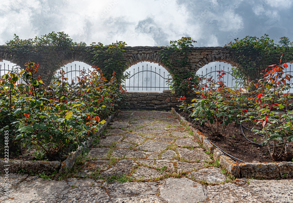 Naklejka premium A paved stone path leads toward a scenic stone wall with arches, wrought iron fencing, and blooming rose bushes under a cloudy sky.