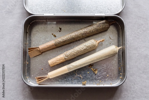 Three pre-rolled cannabis joints, some with paper crutches, inside an open silver tin container, against a light gray textile background.