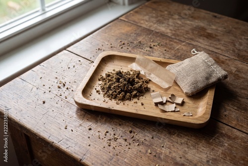 Dried botanical material, rolling papers, and a burlap pouch arranged on a rustic wooden tray near a sunlit window.