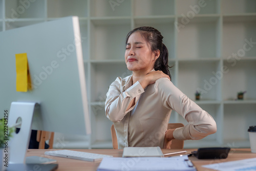 Young Asian businesswoman sitting at her office desk experiencing pain and discomfort in her shoulder and lower back