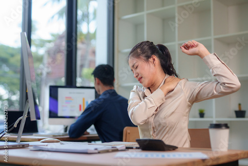 Young Asian businesswoman stretching her stiff shoulder, experiencing pain and discomfort from long hours working at a desk