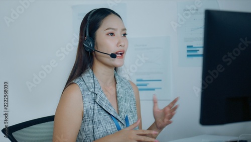 A woman wearing a headset for office communication raises both hands in an explanatory gesture, with the headset microphone supporting her seated interaction beside workplace equipment.