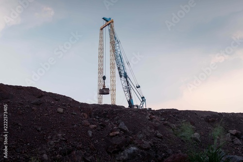 Dynamic compaction crane working on a large earth pile at a construction site against a twilight sky