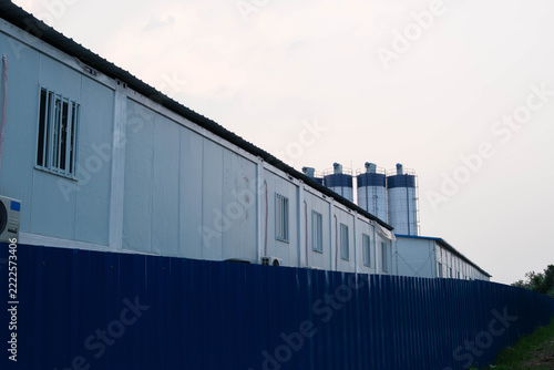 Prefabricated temporary building and blue corrugated metal fence with cement batching plant silos in the background
