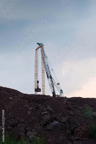 Vertical view of a dynamic compaction crane operating on a rocky earth mound against a twilight sky