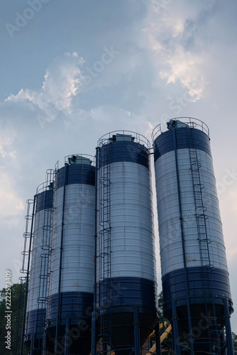 Low angle vertical view of four industrial cement silos at a concrete batching plant against a cloudy sky