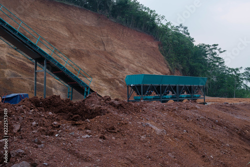 Green aggregate hopper bins and conveyor belt at an earthwork construction site against a terraced dirt slope