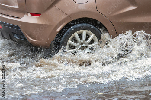 A car navigates through a flooded city street as rain pours down. Water splashes around the wheels, creating puddles and challenges for drivers