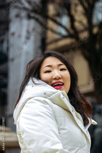 Japanese woman smiling in a white winter jacket outdoors with blurred trees and building behind her, vertical portrait photograph in natural light, Christmas advent 