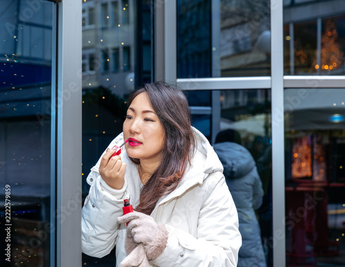 Japanese woman applying red lipstick outdoors while wearing a white winter coat and gloves near reflective glass doors in an urban shopping district, Christmas advent 
