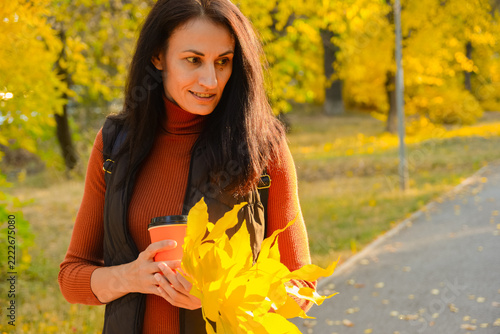 A attractive brunette girl in a warm vest strolling along a path in a beautiful autumn park, holding a paper coffee cup and bright yellow fallen leaves.