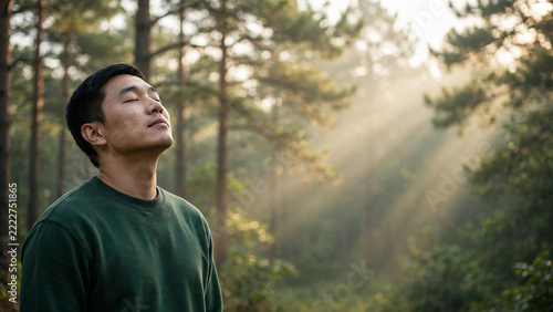 Asian man with closed eyes in sunbeam filled forest perfect for zen meditation mindfulness calm and balanced peaceful living concepts